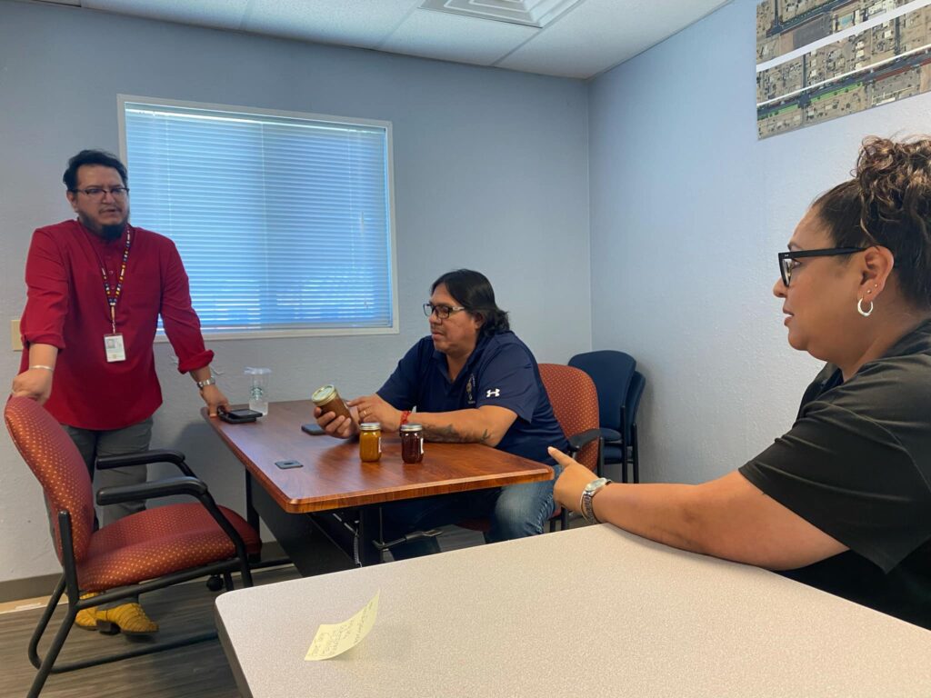 Three people meet inside a Tribal Employment Rights Office; a tribal staff member in a red shirt stands near a chair, another sits examining jars of goods, and a woman entrepreneur sits across the table discussing her application for a TERO business license on the Pascua Yaqui reservation.